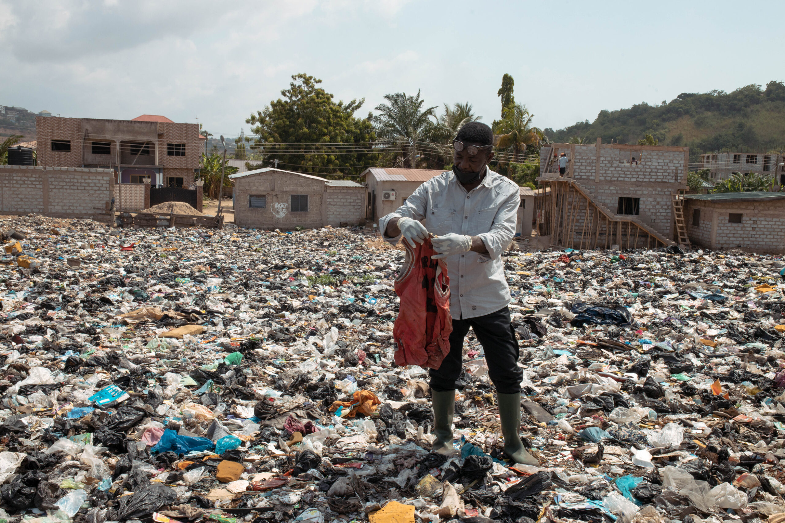 Un homme portant des gants et des bottes examine un vêtement rouge tout en se tenant dans une vaste décharge recouverte de vêtements jetés et de déchets plastiques, entourés de maisons partiellement construites dans un quartier résidentiel.